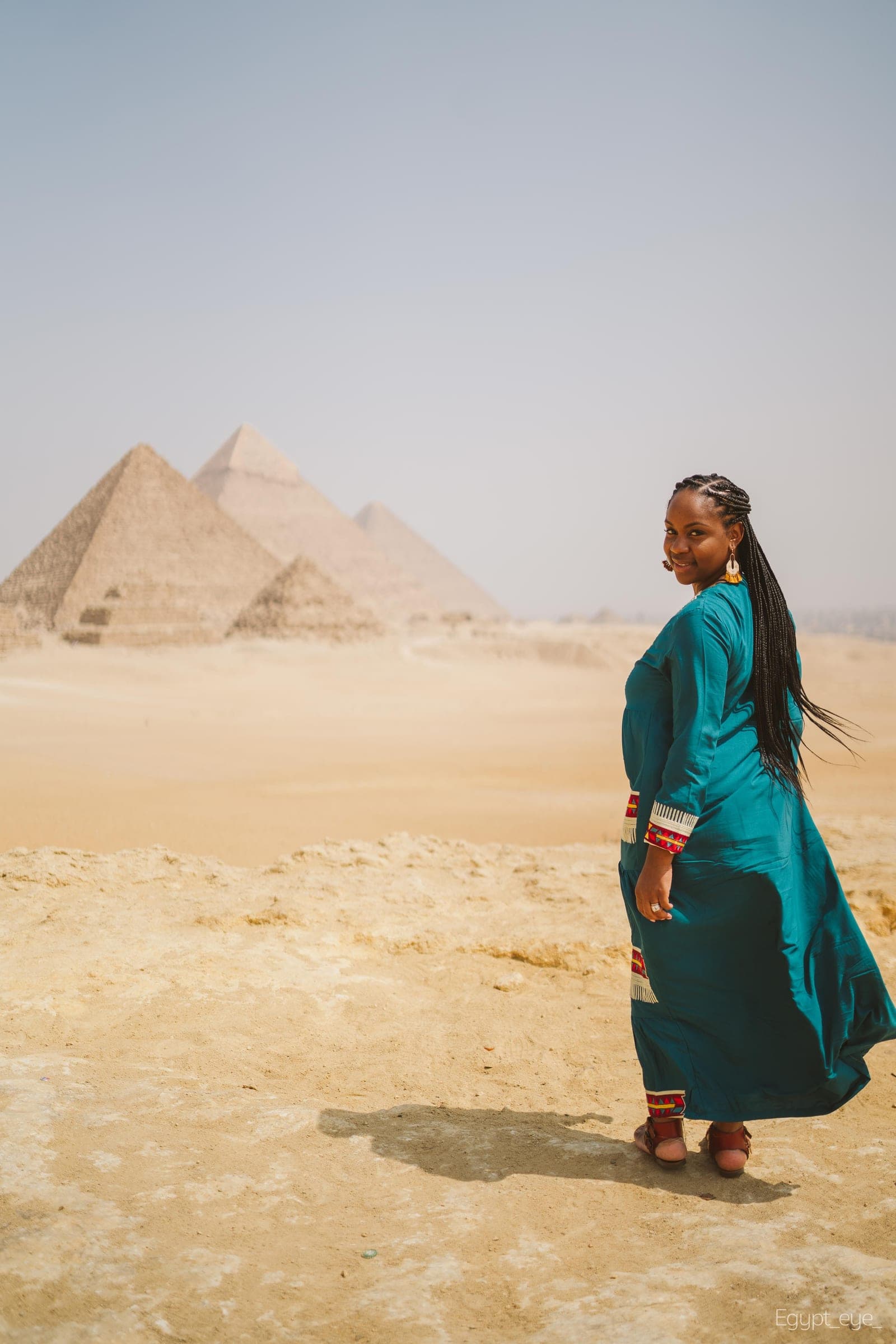 Neké Akua in a teal embroidered kaftan looking back over her shoulder on the sand at Giza, the pyramids behind her.