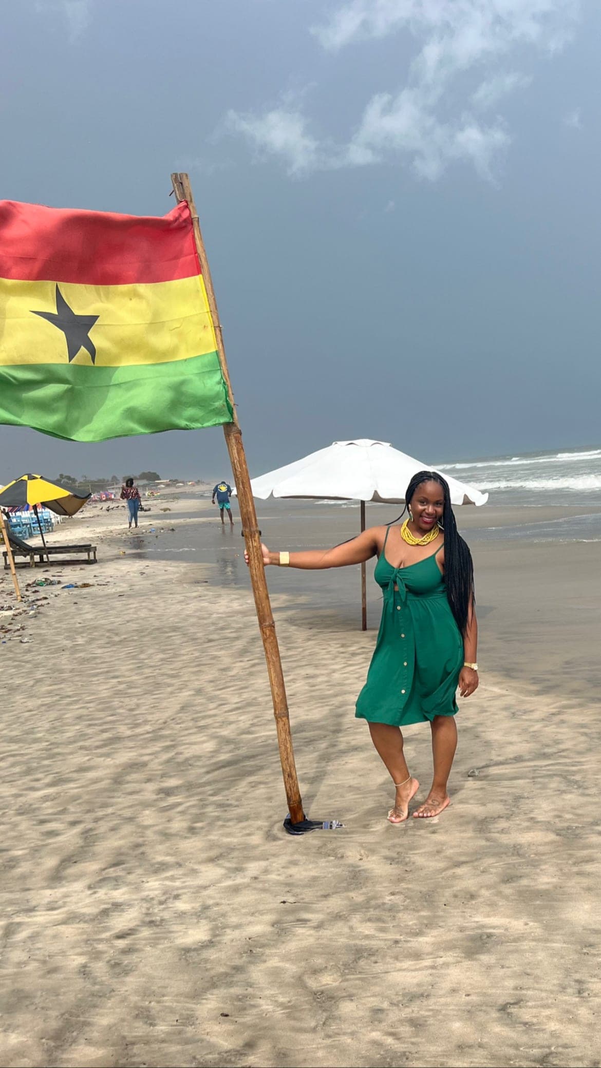 Neké Akua in a green button-front dress, standing on a beach in Accra beside a large Ghanaian flag, ocean waves behind her.