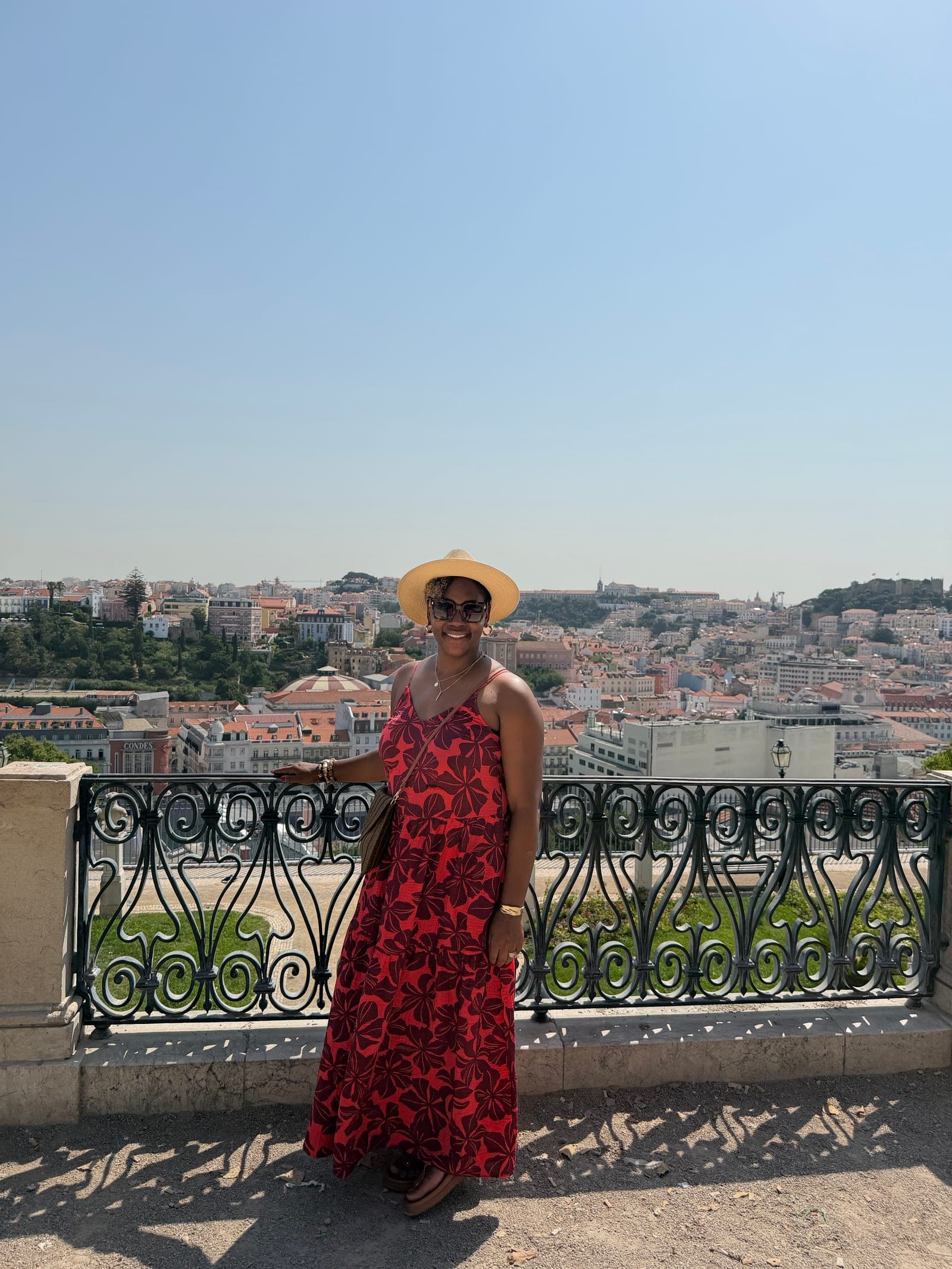 Neké Akua in a red floral maxi dress and straw hat, leaning against a wrought-iron railing overlooking Lisbon’s red-tiled rooftops.