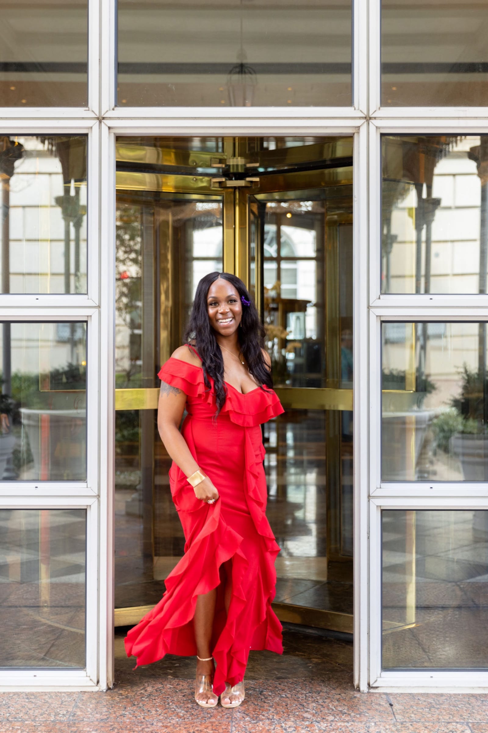 Neké Akua in a red ruffled off-the-shoulder gown, standing in the gold revolving door of a hotel entrance, the train of the dress spread across the stone at her feet.
