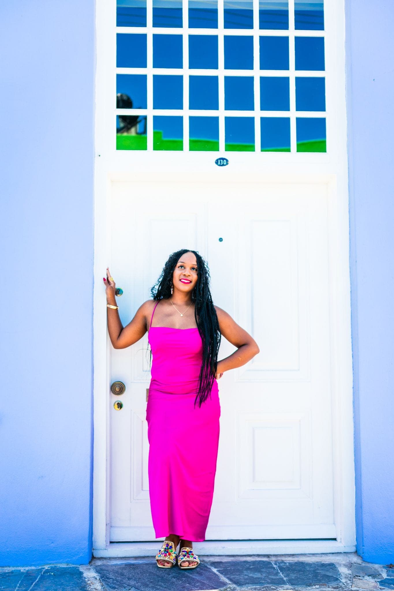 Neké Akua in a fuchsia slip dress at a white Bo-Kaap door with blue and green stained-glass panes, the house painted cornflower blue around it.