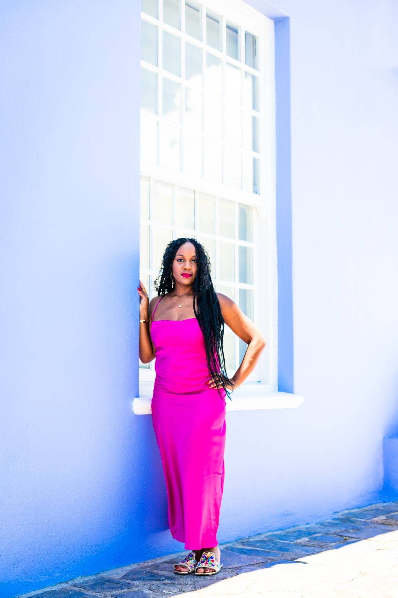 Neké Akua in a fuchsia slip dress beside a white sash window set into a periwinkle-blue Bo-Kaap wall.
