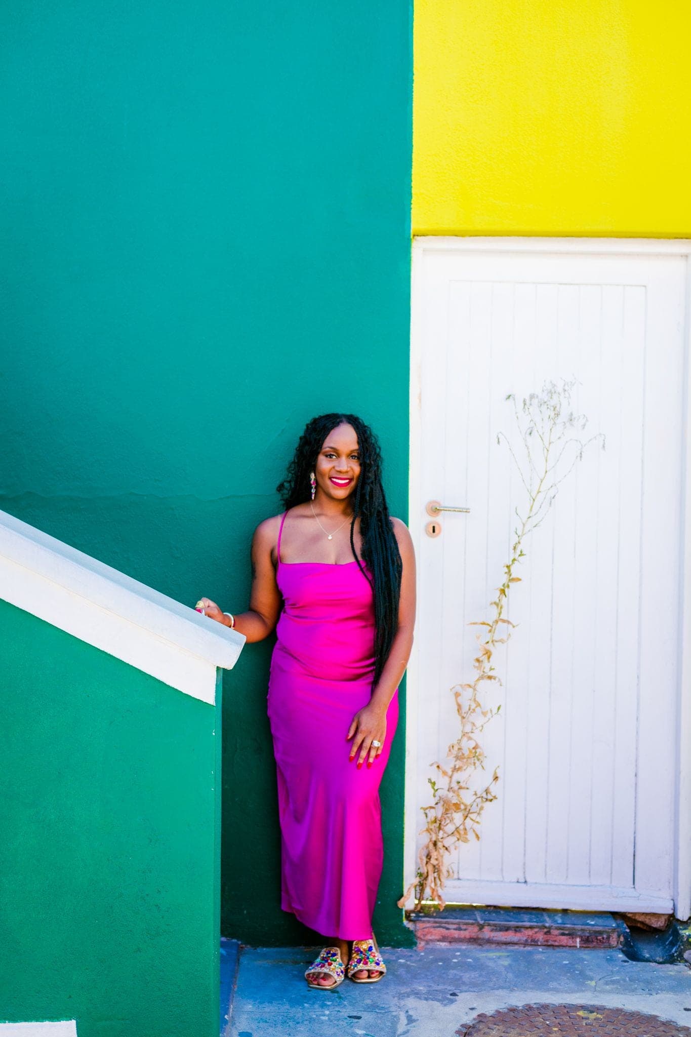 Neké Akua in a fuchsia slip dress by the white steps of a teal Bo-Kaap house, a yellow wall on the corner beyond.