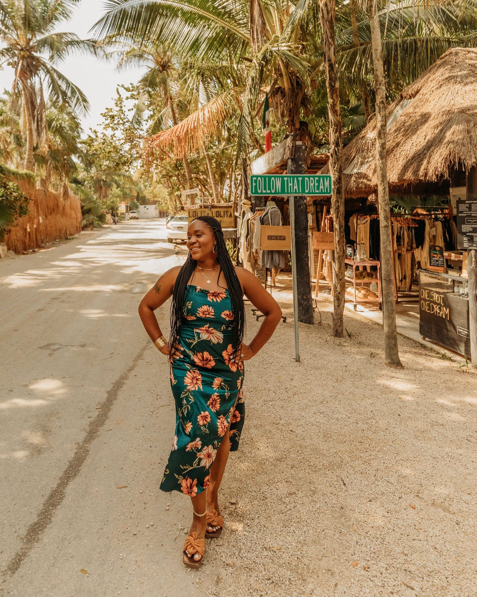Neké Akua standing under Tulum's green FOLLOW THAT DREAM street sign in a floral satin dress, hands on her hips.