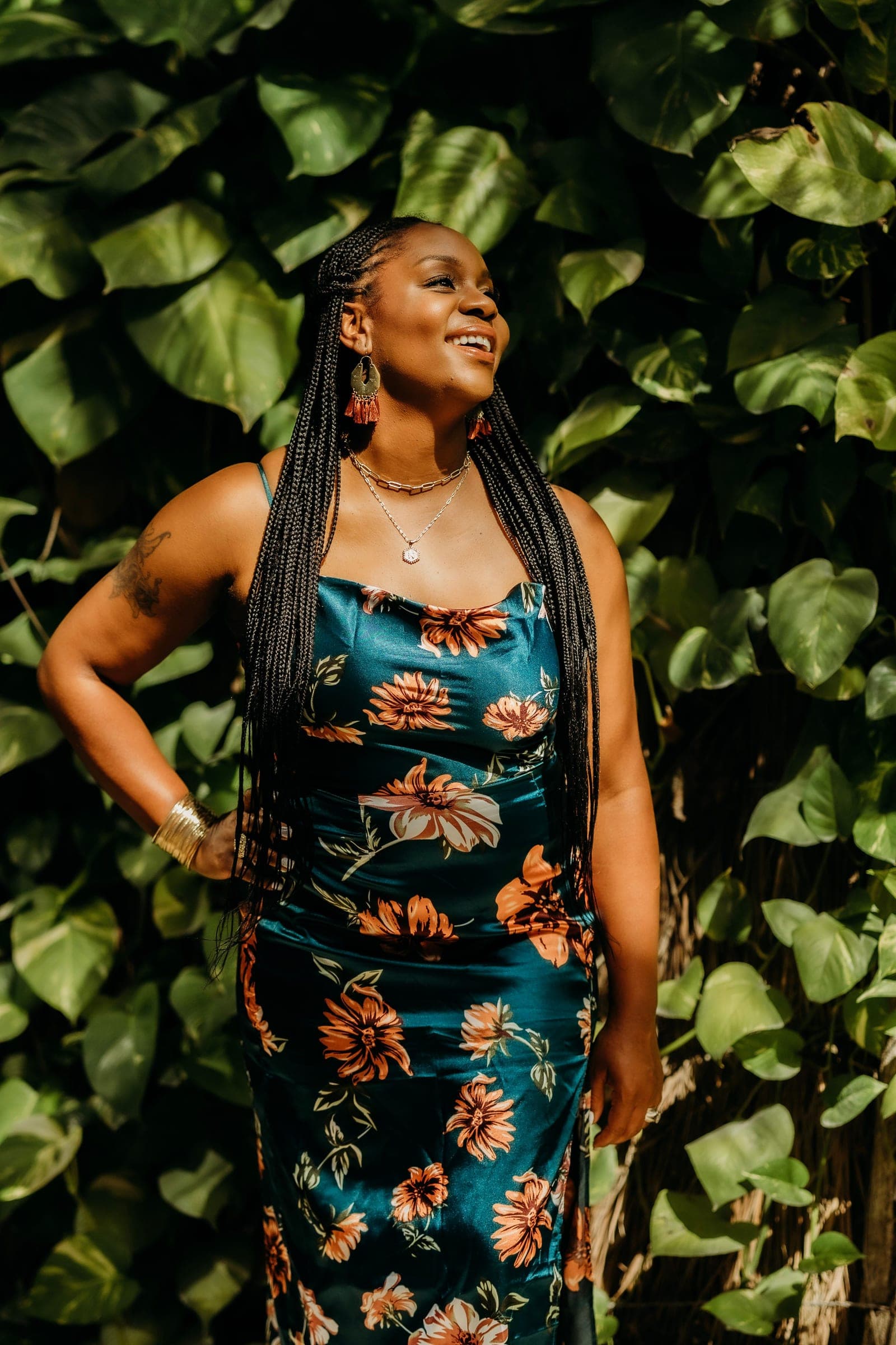 Neké Akua in a teal floral satin dress, hand on her hip, laughing against a wall of glossy green leaves in Tulum.