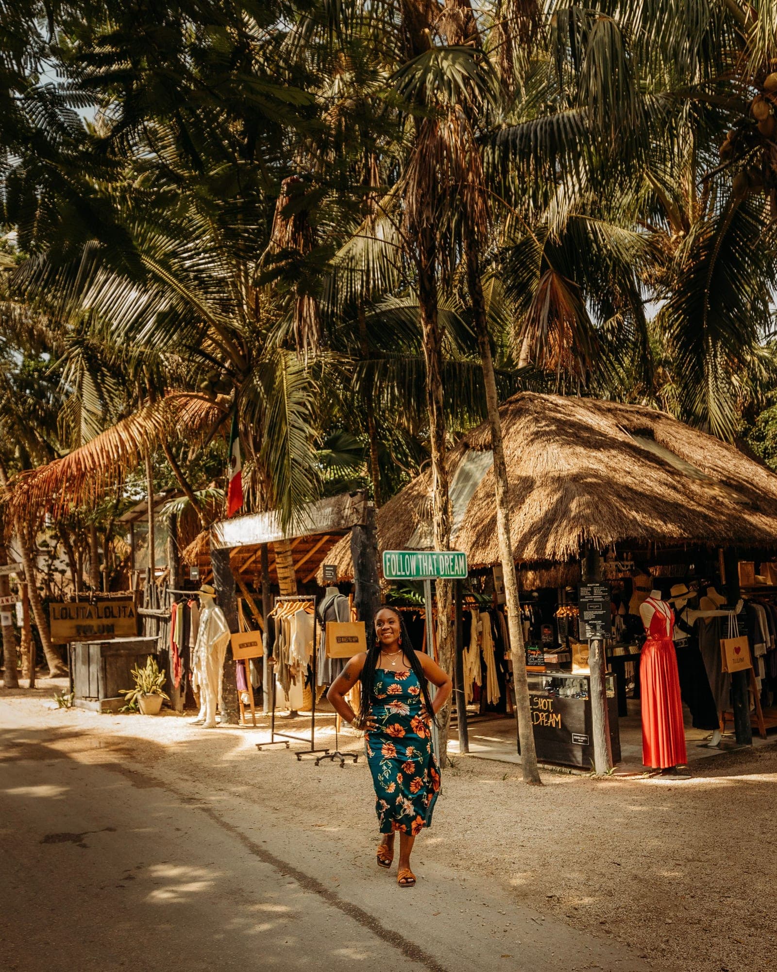 Neké Akua walking a sandy Tulum street in a floral satin dress, thatched-roof shopfronts and palms rising on both sides.