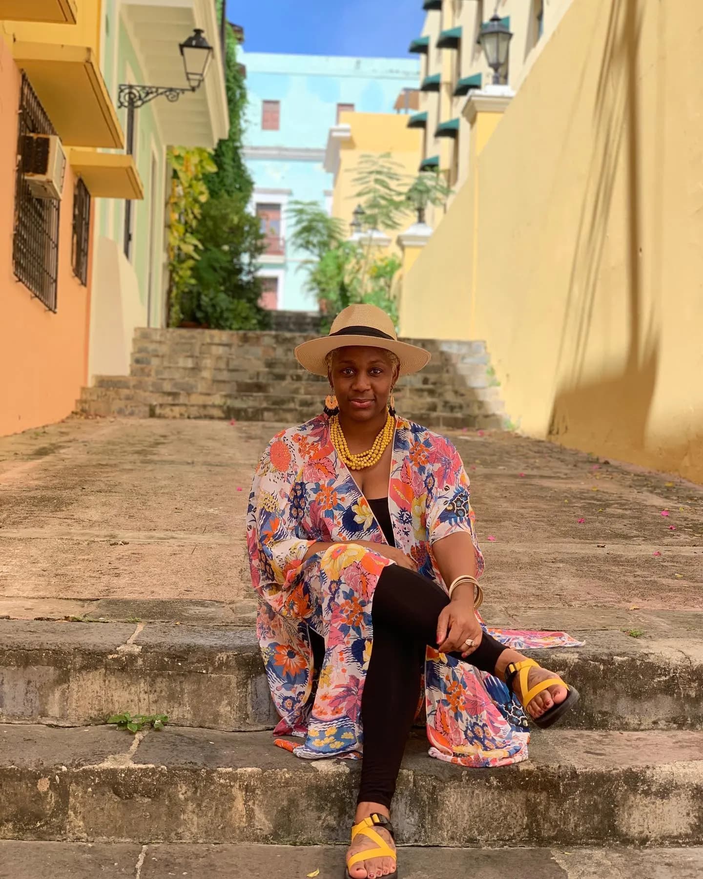 Neké Akua in a floral kimono and sun hat, seated on stone steps between colorful buildings in Old San Juan.