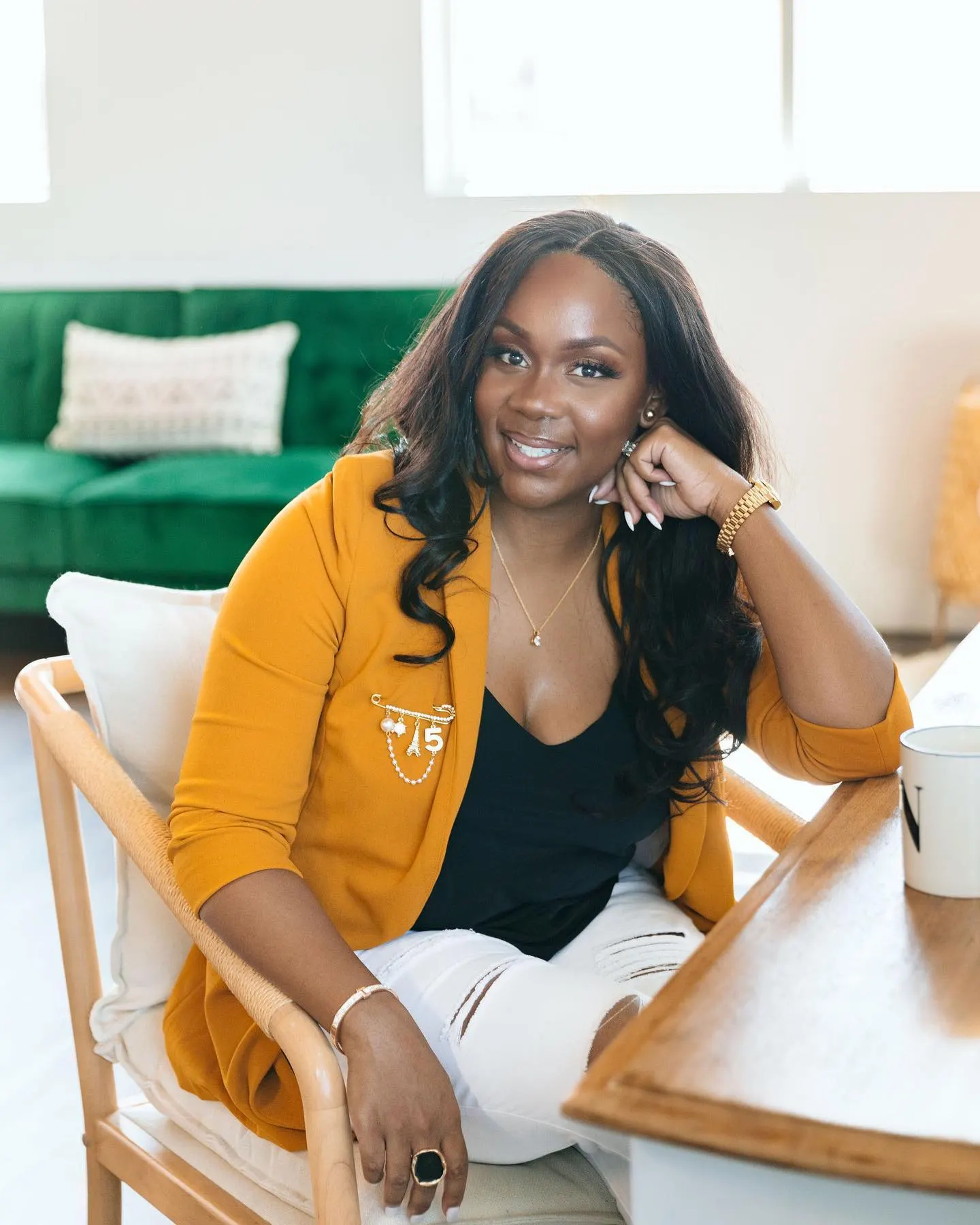 Neké Akua in a mustard blazer with a charm pin, seated in a rattan chair with a green velvet couch behind her.