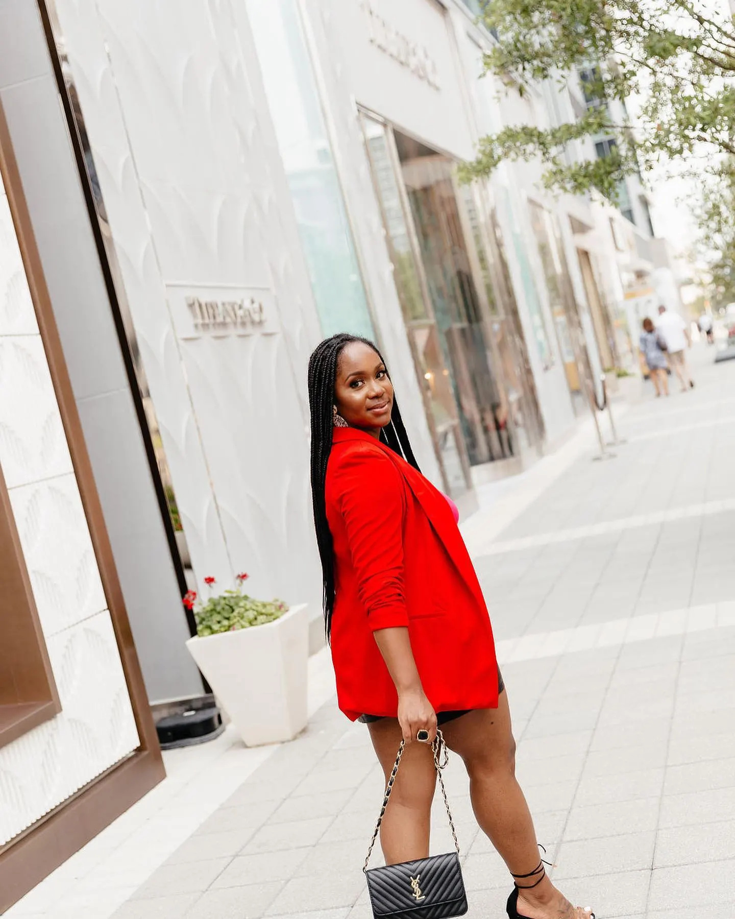 Neké Akua in an oversized red blazer and long braids, glancing back over her shoulder on a sunlit shopping street.