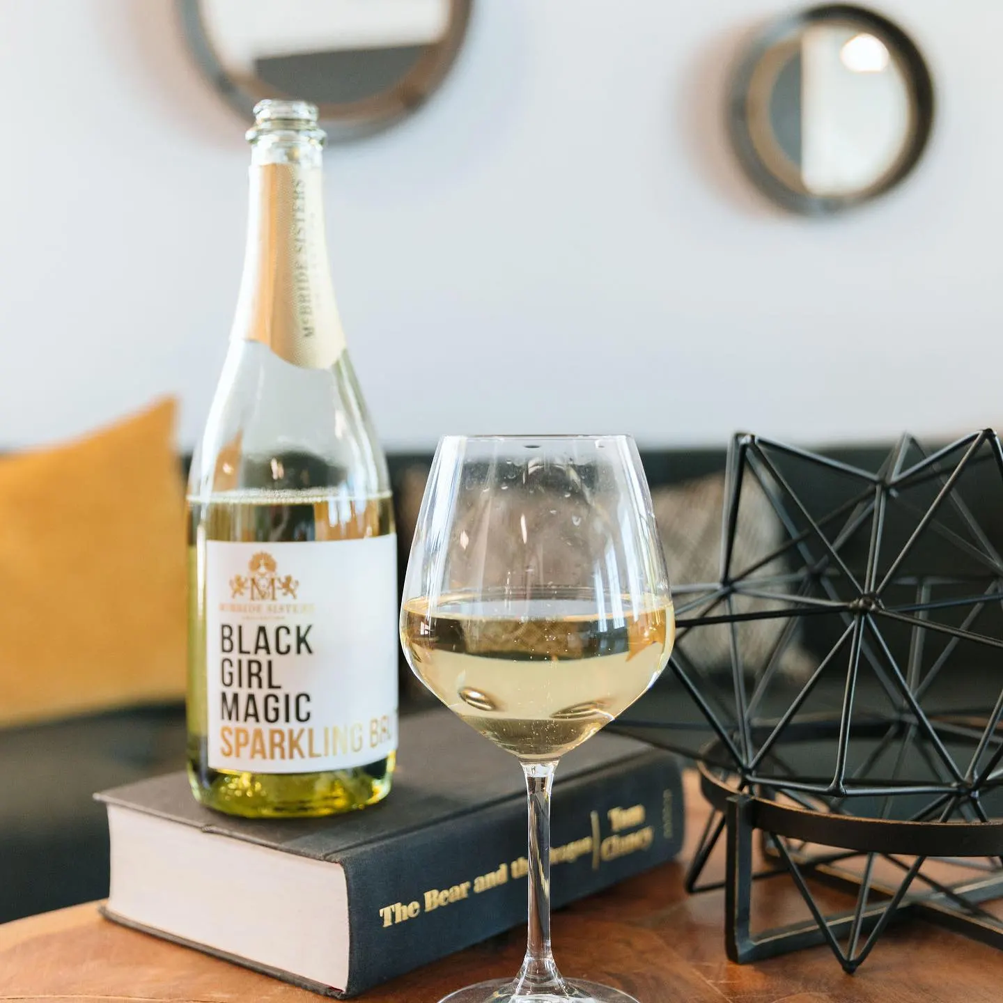 A bottle of Black Girl Magic sparkling wine and a full glass resting on a book, beside a mustard pillow.